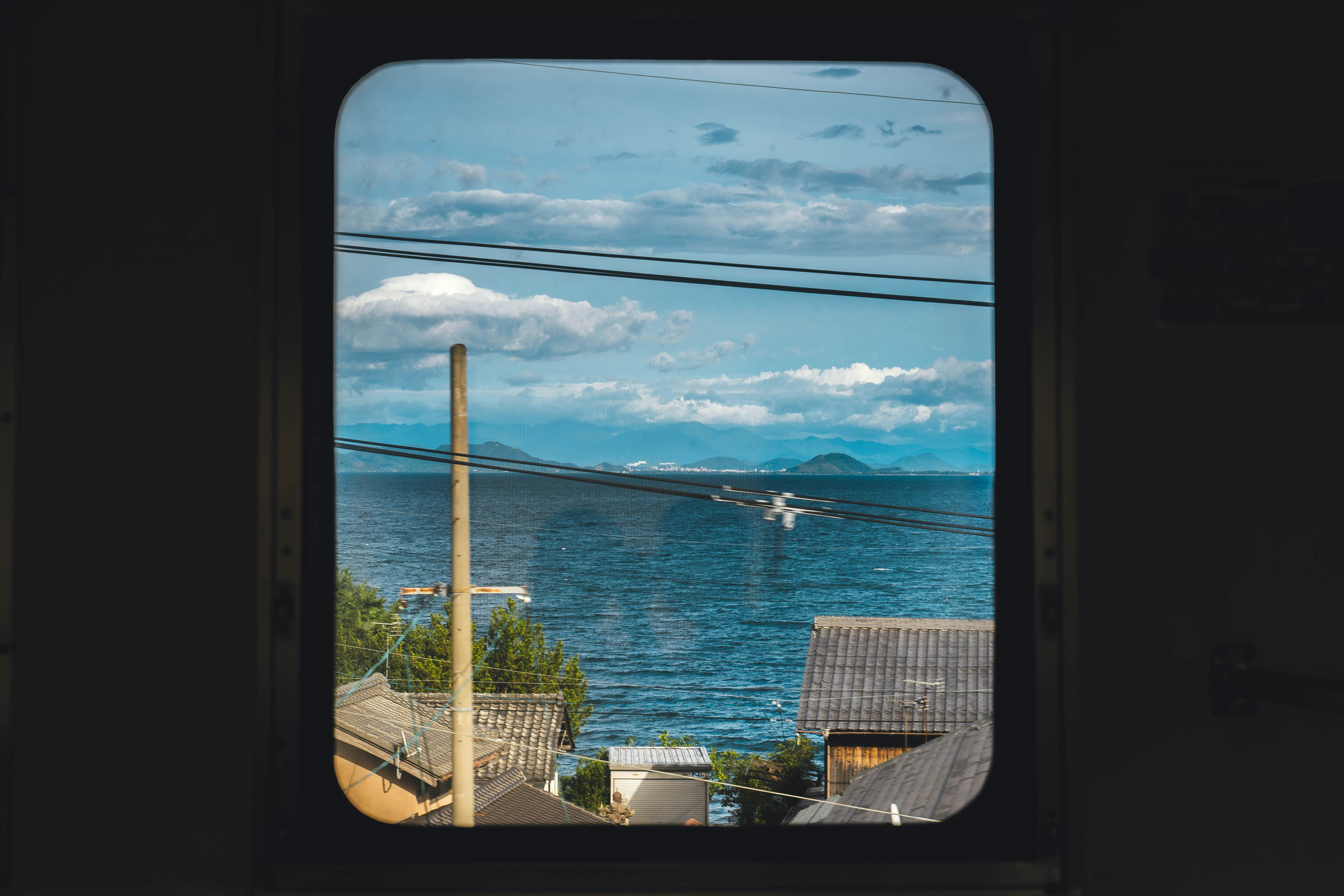 A train window framing a view of open sea, rooftops, and distant hills.